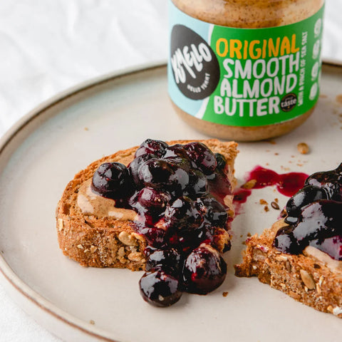 A jar of Yumello Original Smooth Almond Butter sits on a ceramic plate behind two slices of toast topped with almond butter and dark berry jam.