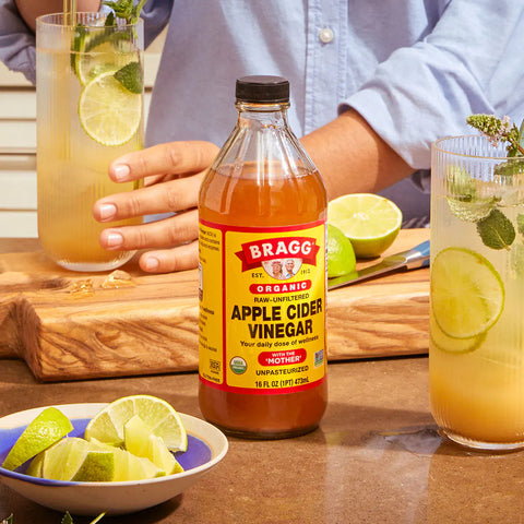 Bottle of Bragg Organic Apple Cider Vinegar on a table with glasses of lemonade and limes.
