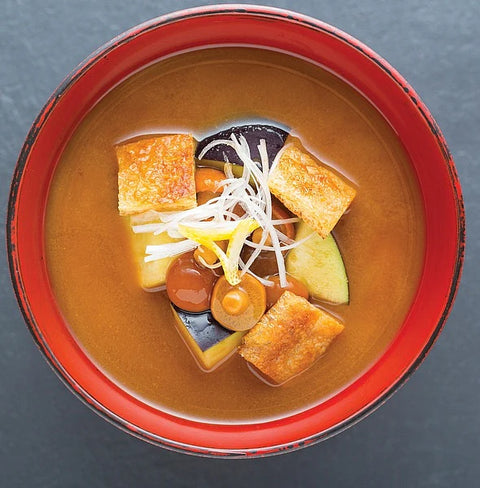 A top-down view of a bowl of Japanese miso soup with tofu, mushrooms, green beans, and shredded white garnish, set against a dark slate background.