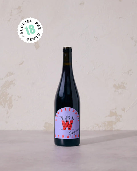 A dark glass bottle of wine with a colorful label, standing on a light surface against a textured off-white wall. A circular badge in the upper left reads 'CALORIES PER CLASS 18'.