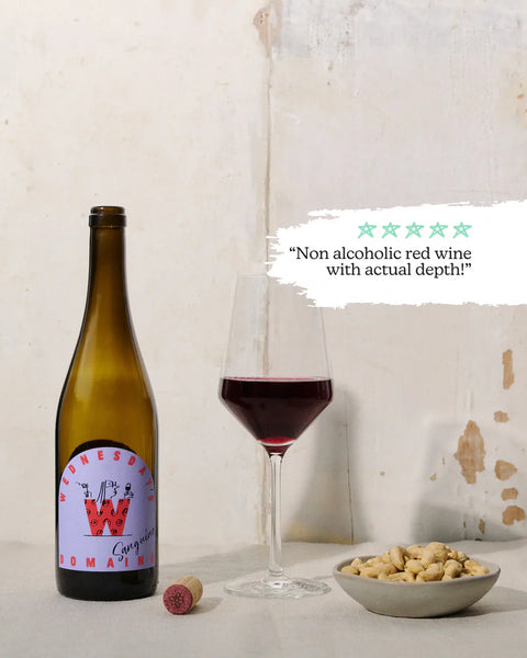 A bottle of non-alcoholic red wine with a cork next to a glass of red wine and a bowl of peanuts, set against a rustic off-white wall with a text overlay.