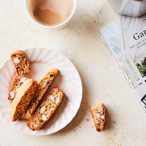 Slices of almond biscotti cookies with sliced almonds and dried fruit, arranged on a white plate with a cup of coffee and an open magazine on a light beige surface.