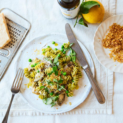 A styled overhead food scene showing a plate of pasta salad with green peas, microgreens, and crumbled cheese, alongside a knife, fork, lemon, grater, and crumbs on a white tablecloth.