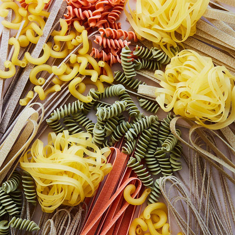 An overhead close-up arrangement of assorted dried pasta shapes in yellow, green, orange-red, and beige on a neutral surface.
