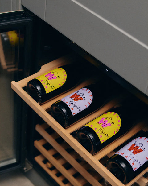 A close-up of a wine cooler's wooden pull-out shelf holding several dark wine bottles with colorful labels, including one with a yellow label and another with a white label featuring a red 'W' logo.