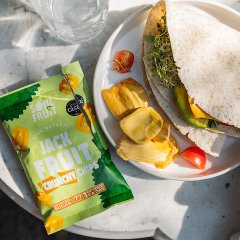 Packaged jackfruit chips next to a sandwich on a plate with a glass of water.