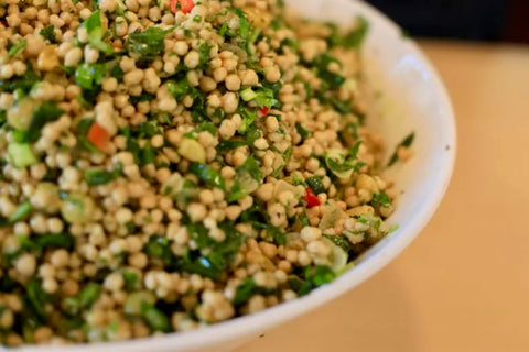 A close-up shot of a white bowl filled with a fresh green and beige grain salad, featuring small beige grains mixed with chopped green herbs and small red pepper pieces.