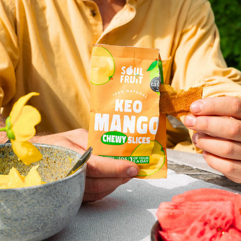 Person holding a packet of 'Kéo Mango' chewy slices with a bowl of chips and fruit in the background.