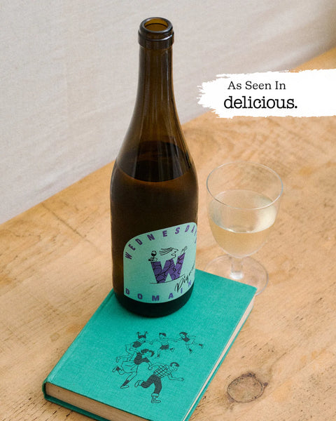 An amber glass bottle of Wednesday Dom Pappy beer stands on a turquoise notebook on a wooden table, next to a filled wine glass. A white text badge in the top right reads 'As seen in delicious.