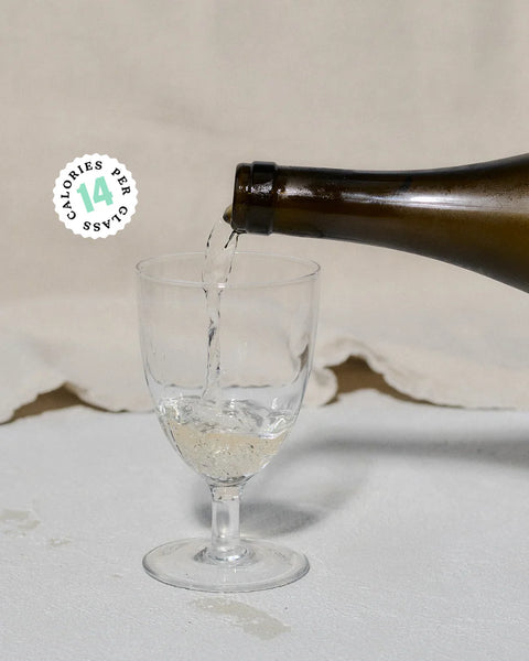 A dark glass bottle pouring a clear bubbly liquid into a clear stemmed glass, set against a neutral beige fabric backdrop. A circular badge on the left reads 'CALORIES PER 14'.