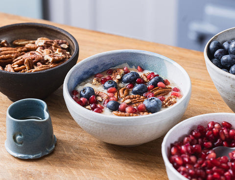 Bowl of cereal with fruit and nuts on a wooden table, surrounded by additional fruits and a small pitcher.