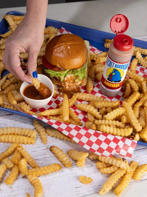 A hand-dipped crinkle-cut french fry meal on a blue tray, featuring a burger, a small cup of sauce, and a bottle of Secret Airdurg Airdurg sauce, surrounded by scattered fries on a white table.