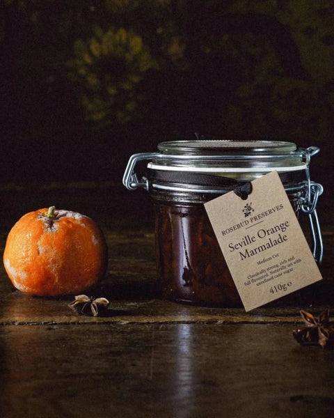 A glass jar of Rosebud Reserves Seville-Orange Marmalade sits on a rustic wooden table next to a fresh orange, with a kraft paper tag hanging from the metal clasp lid.