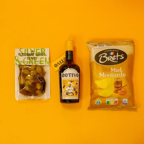Three products on a yellow background: a bottle of Botivo, a bag of Brets mustard chips, and a bag of olives.