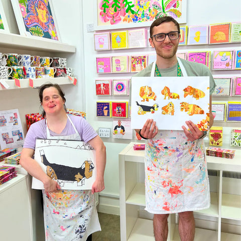 Two people wearing aprons holding up their artwork in a colorful art studio.