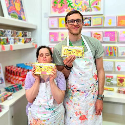 Two people wearing aprons holding boxes in a store with colorful products on shelves.