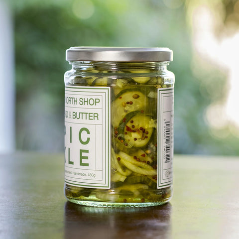 A glass jar of North Shop Pickled & Butter Garlic Chilis on a dark wooden table with a blurred green outdoor background.