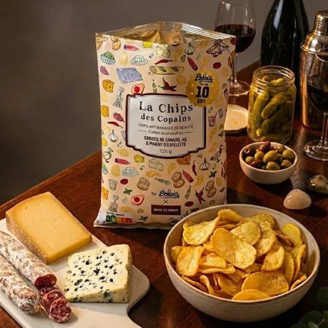 A bag of La Chips des Copains potato chips stands on a wooden table surrounded by food props, including cheese, cured meats, olives, a bowl of chips, and wine glasses and bottles.