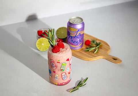 A pink iced cocktail in a clear glass jar-style tumbler with colorful La Croix logo decals, topped with raspberries and a rosemary sprig, alongside a purple La Croix sparkling drink can on a wooden cutting board with lime slices and fresh rosemary.
