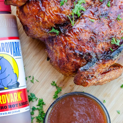 A bottle of BrdVark No. 9ero Hot Sauce next to a large glazed roasted chicken on a wooden board, with a small bowl of sauce and parsley garnish.
