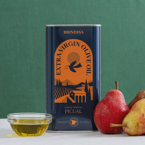 A can of Brindisa Extra Virgin Olive Oil stands on a white surface against a green background, with a small glass bowl of golden olive oil and red and yellow pears placed beside it.