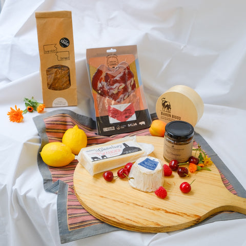 Assorted food items including lemons, cheese, and a jar on a wooden board with branded packages in the background.