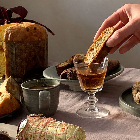 A hand holds a biscotti cookie over a small glass of amber beverage on a table set with bread, a ceramic mug, and plates in a warm, rustic indoor setting.