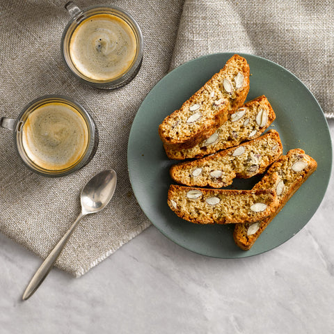 Four slices of sunflower-biscotti cookies on a muted green plate, styled with two cups of espresso and a spoon on a linen napkin.