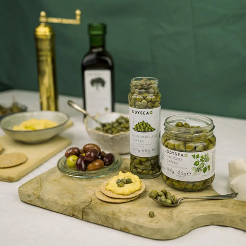 two jars of capers on a wooden cutting board with olives and crackers