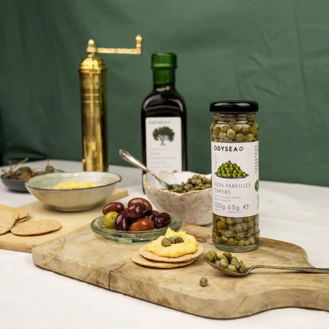jar of capers on a wooden cutting board with crackers and olives