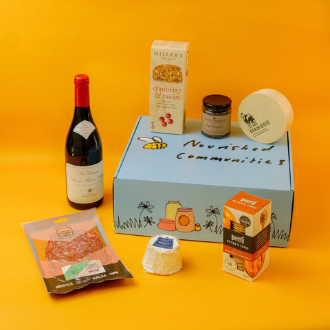 Assorted foods including a bottle of wine, cheese, and packaged items on an orange background with a 'Nourished Communities' hamper box.