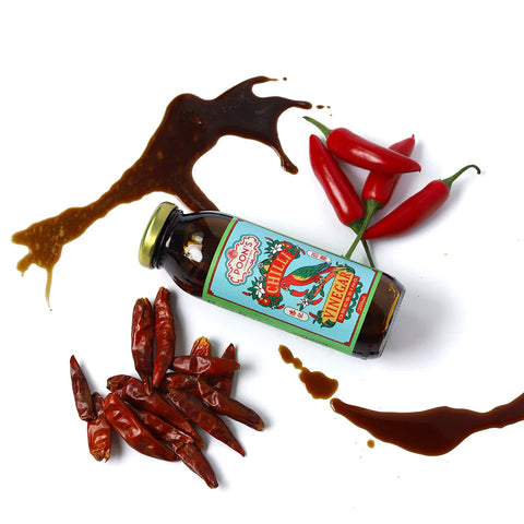 Bottle of hot sauce with a colorful label, surrounded by red chili peppers and dried chili pods on a white background.