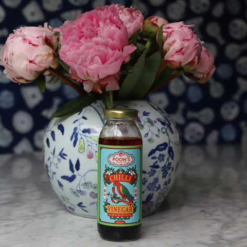 Bottle of Poon's Chilli Vinegar in front of a vase with pink flowers on a marble surface.