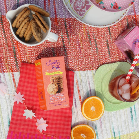A styled flat-lay featuring a box of Sweet Fingers Creamy Orange Cookies, surrounded by cookies in a white mug, orange slices, a pink drink on a green saucer with a striped straw, and a colorful patterned tablecloth.