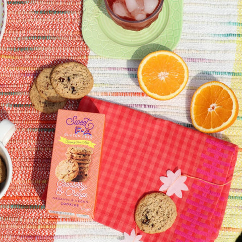 A box of Sweet Fia Gluten Free Vanilla Spice Cookies is styled in a bright, colorful flat-lay with cookies, orange slices, and a glass of iced drink on a striped tablecloth.