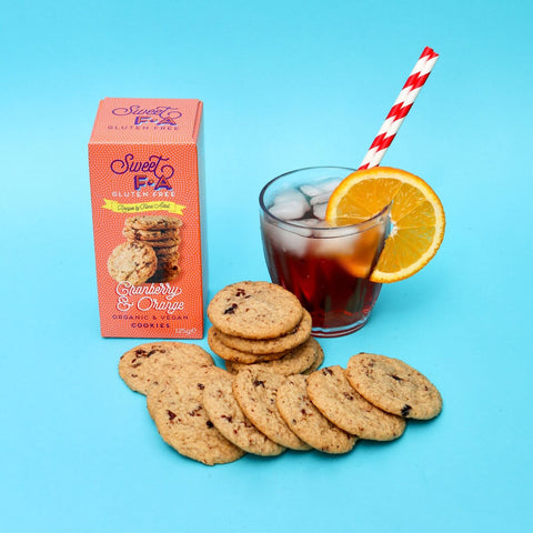 A box of Sweet FA Gluten Free Slightly Orange Cookies sits next to a glass of iced dark cola with an orange slice and a red-and-white striped straw, with several cookies spread in front on a bright blue background.