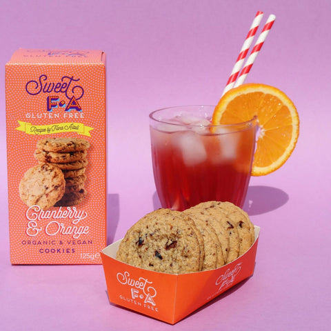 Sweet F&A Cranberry & Orange Gluten Free Cookies shown with their orange packaging box, a tray of cookies, and a glass of iced drink with an orange slice garnish on a pink background.