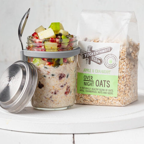 A jar of overnight oats topped with fruit pieces and a spoon, next to a bag of Yockenthwaite Farm Apple & Cranberry Over Night Oats, on a white surface against a white background.