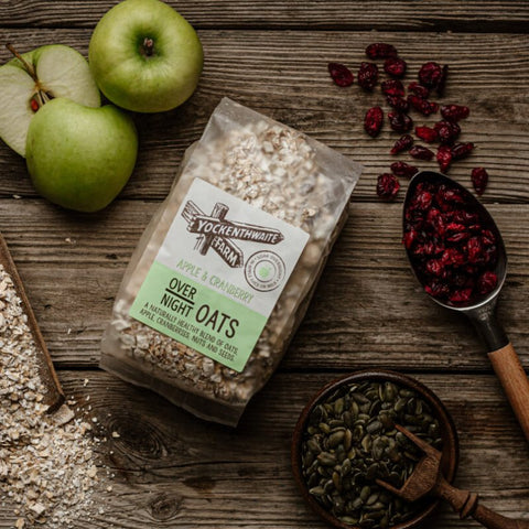 A bag of Tockenwhite Farm Over Night Oats (Apple & Cranberry flavor) is displayed on a rustic wooden table alongside fresh green apples, dried cranberries, pumpkin seeds, and oats.