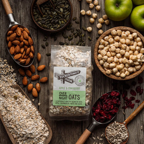 A bag of Over Night Oats (Apple & Cranberry) is displayed on a rustic wooden table surrounded by its raw ingredients, including almonds, green apples, chickpeas, oats, sunflower seeds, and dried cranberries.