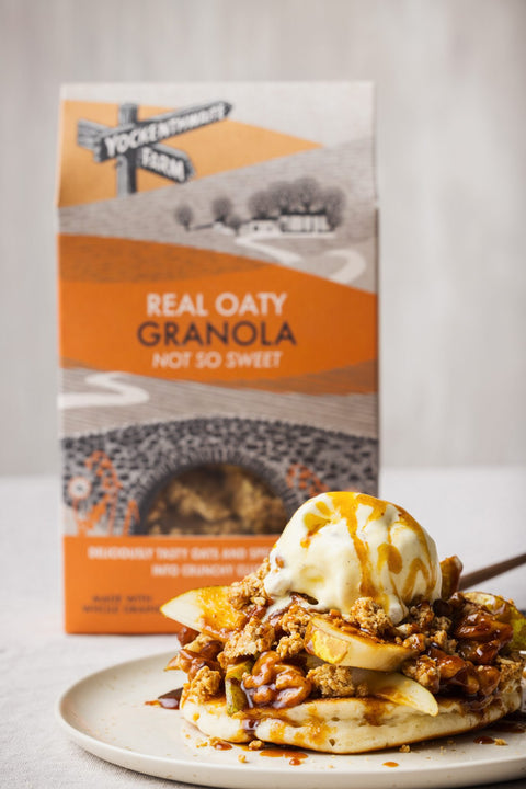 A plate of oatmeal topped with banana slices, granola, and a scoop of vanilla ice cream with drizzled syrup, set in front of a box of Yockenthwaite Farm Real Oaty Granola.