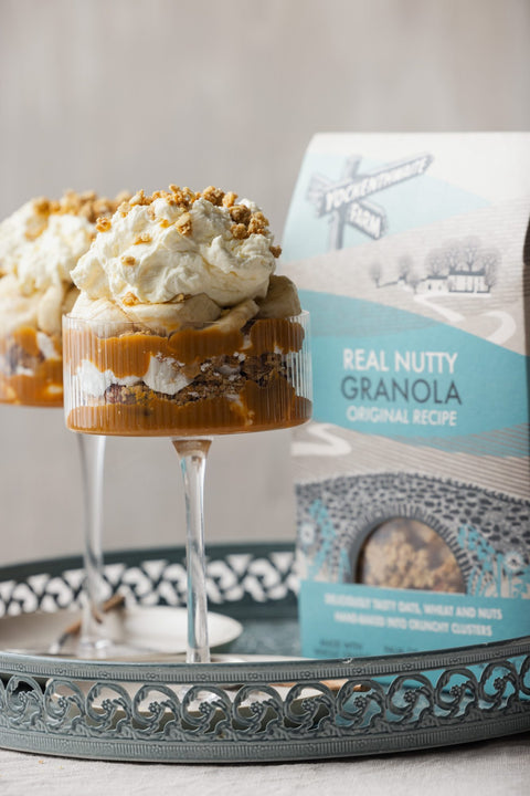Two dessert parfaits in clear glasses on a decorative tray, with a bag of Yockenthwaite Real Nutty Granola Original Recipe standing behind them against a soft neutral background.