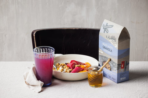 A bag of Granola sits on a table next to a glass of purple drink, a bowl of muesli with fruit, and a jar of honey with a wooden dipper, set against a dark chair and textured light gray wall.
