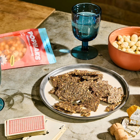 A styled snack scene on a stone tabletop with a plate of textured brown jerky or dried meat slices, a blue goblet, a bowl of peanuts, and a bag labeled Poppa's in the background.