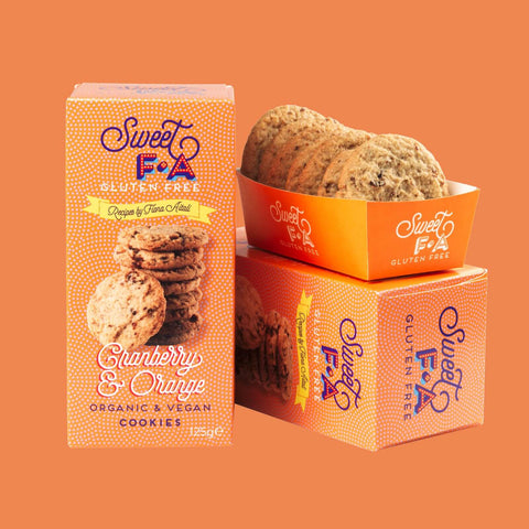 Sweet F.A Gluten Free Cranberry & Orange cookies shown with their orange packaging boxes and a tray of cookies on a solid orange background.