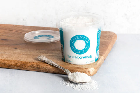 Cornish Sea Salt crystals container on a wooden board with a spoonful of salt.