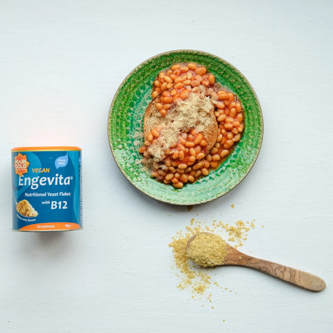 A can of Enevita Vegan Nutritional Yeast with B12 next to a green ceramic bowl containing cooked baked beans with a piece of toast and a wooden spoon of loose yellow powder on a white background.