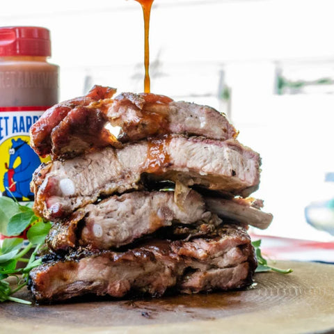 A close-up of a stacked pile of grilled pork ribs with a glossy sauce being drizzled over them, garnished with green herbs, with a bottle of barbecue sauce in the blurred background.