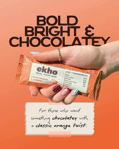 A hand holds an Ekho Real Food Bar wrapped in orange and white packaging against a peach-orange background with bold text reading 'BOLD BRIGHT & CHOCOLATEY' and a note below.