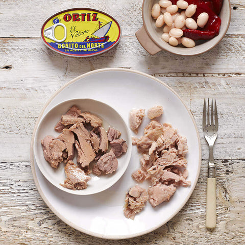 Diced meat on a plate with a can of Ortiz tuna and a bowl of beans and peppers on a wooden surface.